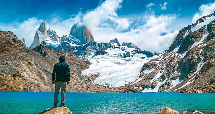 Ein einsamer Wanderer steht auf einem Felsen und blickt auf eine türkisfarbene Lagune und die majestätischen schneebedeckten Gipfel des Fitz-Roy-Massivs unter einem strahlend blauen Himmel.