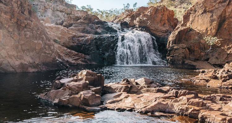A clear waterfall tumbles over dark rocks into a tranquil pool framed by rugged, reddish cliffs and sparse trees.