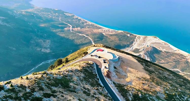 Vue aérienne spectaculaire d'une route côtière sinueuse, d'une plateforme de point de vue et d'une mer turquoise scintillante loin en dessous des crêtes de montagne.