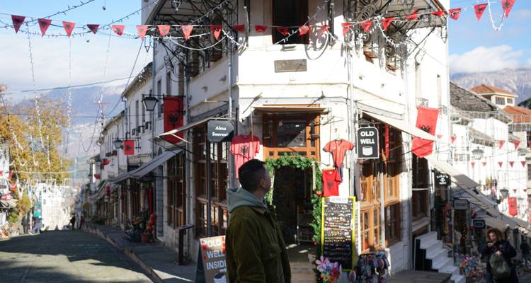Rue du bazar historique avec des bâtiments blancs traditionnels décorés de drapeaux albanais rouges, un visiteur solitaire au premier plan.