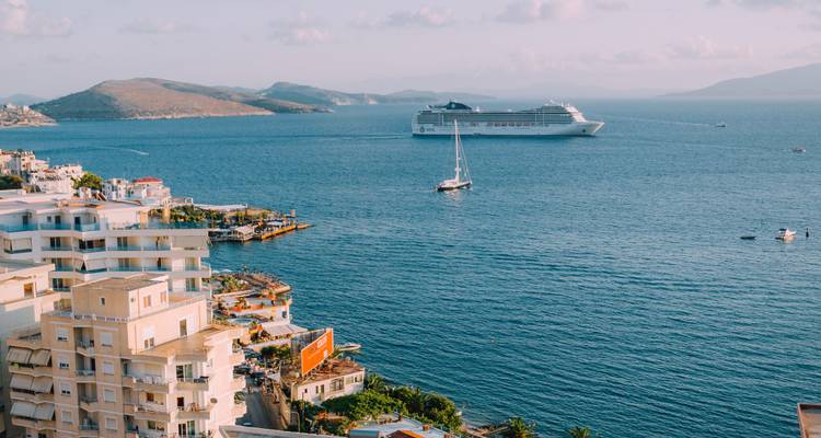 Baie bleue avec un navire de croisière et un voilier mouillés au large d'une ville côtière albanaise aux immeubles d'appartements blancs.
