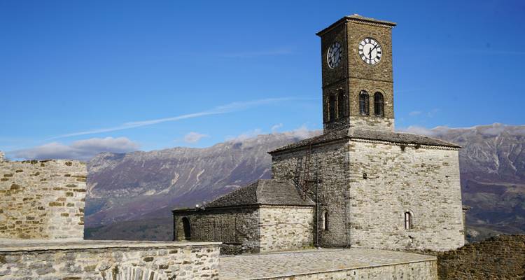 Tour d'horloge en pierre de la forteresse de Gjirokastër se dressant sur un ciel bleu dégagé et une chaîne de montagnes escarpées.