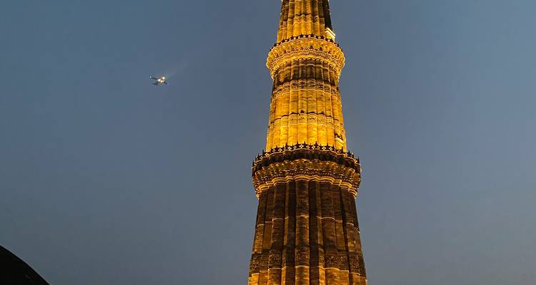 Illuminated Qutub Minar soaring into dusk sky with a passing airplane nearby.