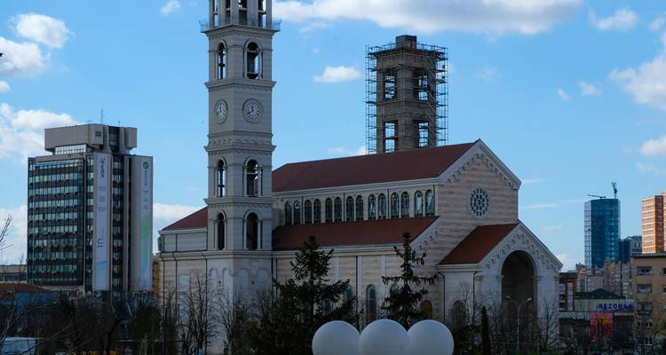 Catedral parcialmente construida con torres gemelas y andamios de pie entre bloques modernos de oficinas bajo un cielo azul.