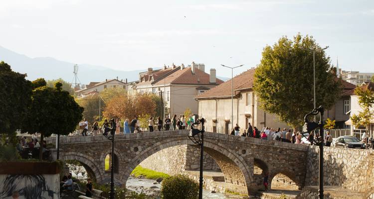 Pont en pierre aux arches historiques à Prizren avec de nombreux piétons profitant d'un après-midi ensoleillé.