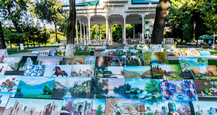 Cuadros de paisajes coloridos se alinean en un mercado de arte al aire libre bajo árboles frondosos en un parque de la ciudad.