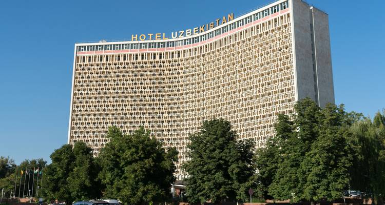 La fachada modernista curva del Hotel Uzbekistán se alza sobre árboles frondosos bajo un cielo azul despejado.