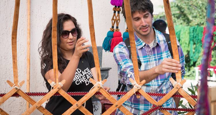 Dos viajeros aprendiendo técnicas de tejido tradicional en un telar de madera al aire libre.