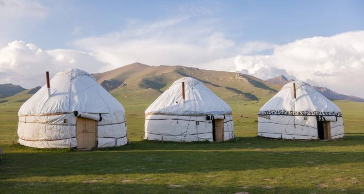 Drie traditionele witte yurts staan op een breed groen steppegebied omringd door glooiende heuvels onder een blauwe hemel met verspreide wolken