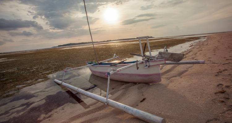 Ein traditionelles Auslegerkanu ruht bei Ebbe an einem ruhigen Strand unter einem warmen Sonnenuntergang.