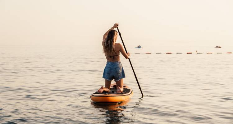 Young woman stand-up paddleboarding alone on calm sea water under hazy sky