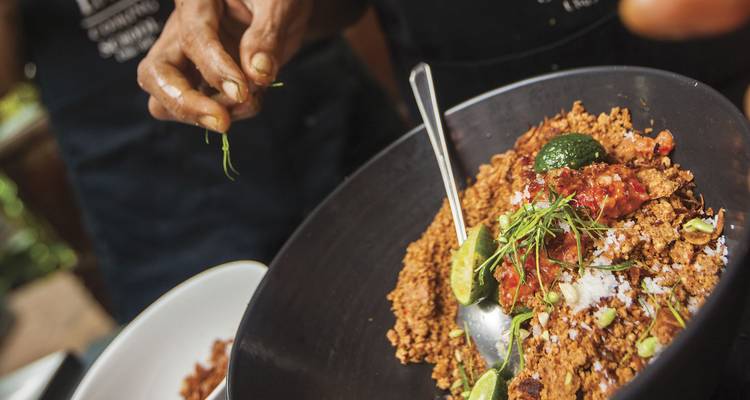 Chef’s hand sprinkling herbs over a traditional Balinese dish in a bowl.