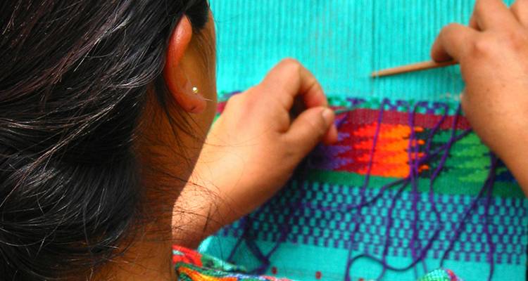Artesano tejiendo un textil colorido en un telar de cintura tradicional, visto desde atrás del hombro.