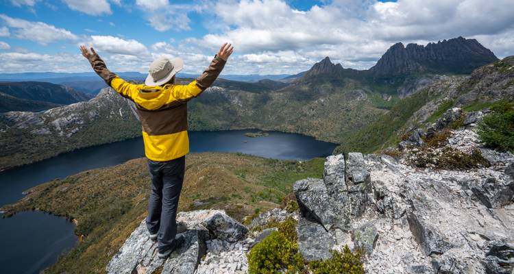 Persoon die op een rotsachtige klif staat met opgeheven armen, uitkijkend over een schilderachtig bergmeer.