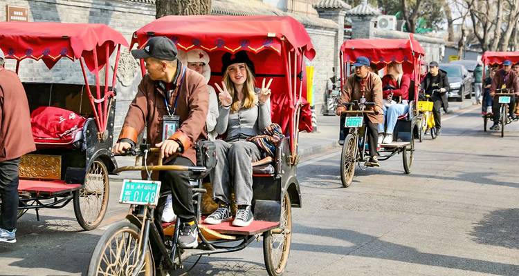 Turistas montando en rickshaws a través de un callejón hutong histórico en Pekín, sonriendo y saludando con la mano.