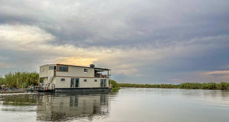 Houseboat floating on a calm river under a cloudy sky.