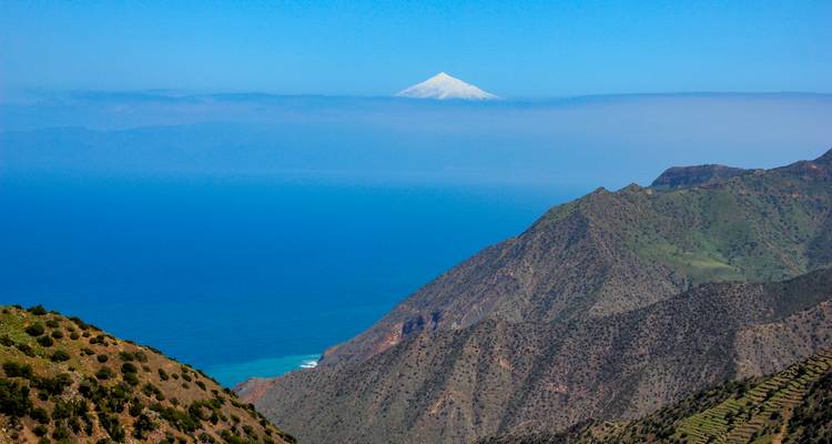 El nevado Monte Teide asoma sobre un mar de nubes más allá de empinadas crestas verdes y el horizonte del Atlántico.
