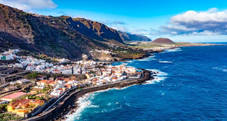 Wide aerial of a rugged coastline road hugging cliffs leading to a seaside town with dramatic mountains beyond.