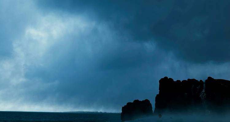 Paisaje marino oscuro y melancólico con nubes de tormenta sobre una costa distante.