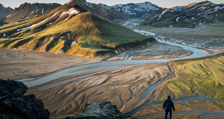 Un excursionista contemplando un vasto y dramático paisaje lleno de ríos y montañas cubiertas de musgo.