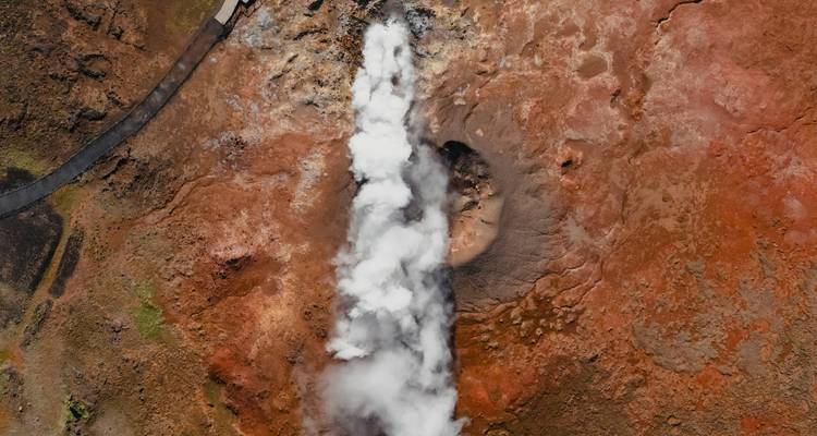 Aerial view of a steaming geyser surrounded by red terrain.