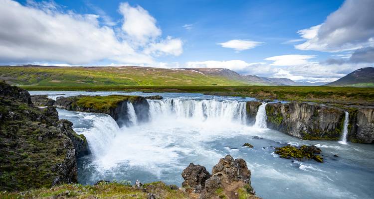 Wide waterfall surrounded by rocky landscape and hills.