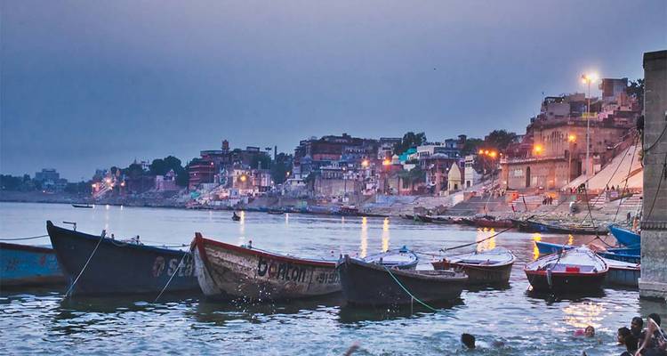 Ganges riverbank with boats and historic buildings at sunset.