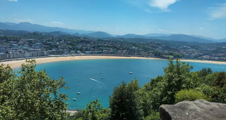 Vue aérienne panoramique de la baie turquoise en croissant et du paysage urbain en bord de mer de Saint-Sébastien bordé de collines verdoyantes.
