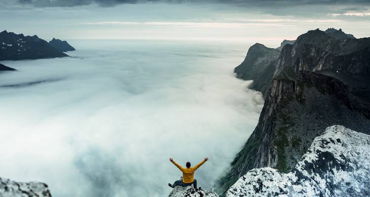 Personne assise au bord d'une falaise avec une mer de nuages en contrebas.
