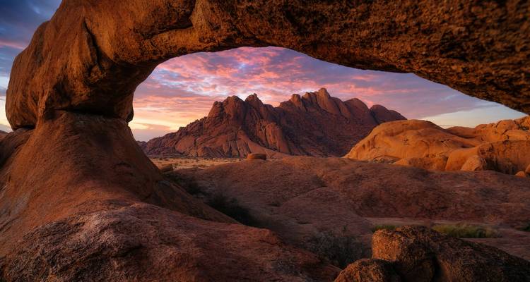 Formations rocheuses sous un coucher de soleil dramatique vues à travers une arche naturelle.