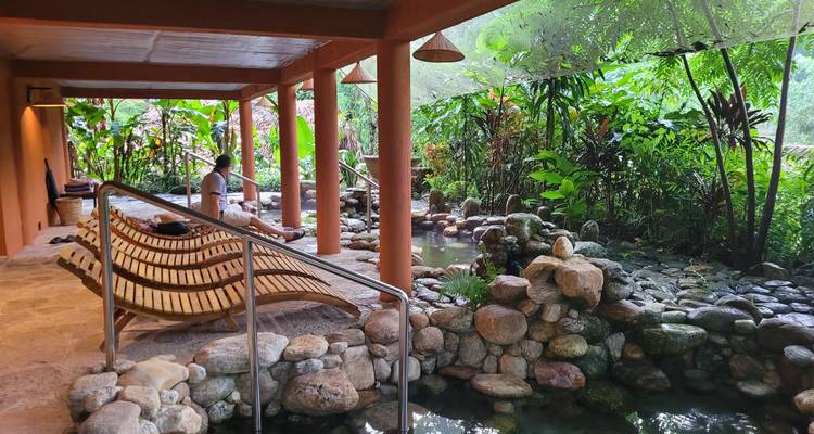 Serene spa corridor with bamboo loungers beside a natural stone hot-spring pool surrounded by jungle plants.