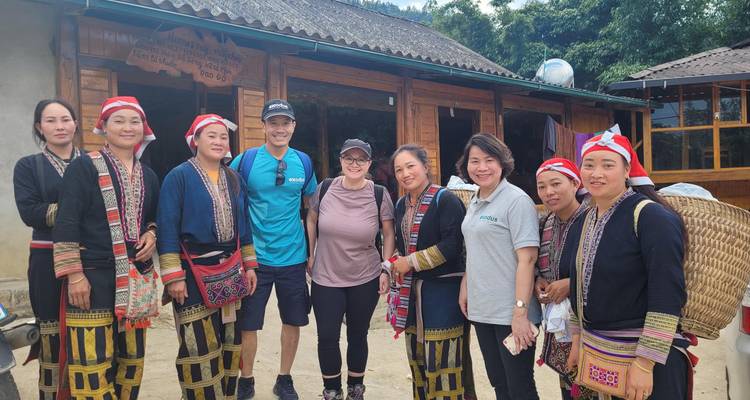 Travellers pose with Red Dao women in traditional embroidered dress outside a wooden house.