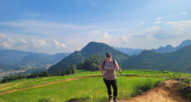 Hiker smiles while trekking through emerald rice terraces with dramatic limestone peaks behind.