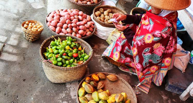 Vendor in conical hat sells baskets of colourful peppers and potatoes at a Vietnamese market.