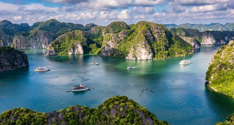 Vue aérienne panoramique d'îles calcaires émeraude parsemées de bateaux de croisière sur des eaux turquoise sous un ciel dramatique.
