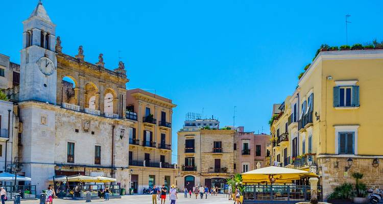 Plaza soleada rodeada de edificios históricos y una torre de reloj en Bari llena de visitantes paseando