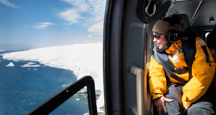 Passager vêtu d'une veste d'expédition aux couleurs vives observe depuis un hélicoptère un littoral antarctique spectaculaire et glacé.