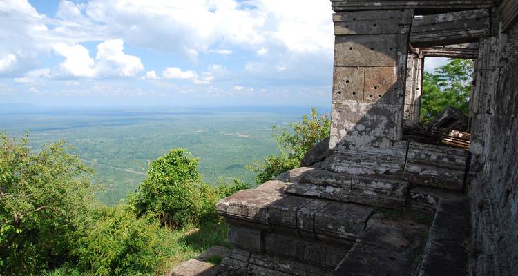 Terraza de piedra de un antiguo templo jmer en lo alto de una colina que domina amplias llanuras de bosque verde bajo un cielo azul brillante con nubes esponjosas.