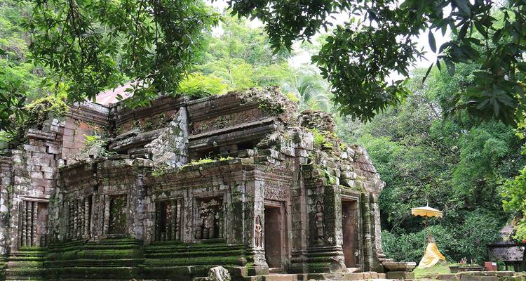 Oude, mosbegroeid Khmer-stijltempel ruïnes omgeven door dicht oerwoud met een eenzame bezoeker die een parasol vasthoudt.