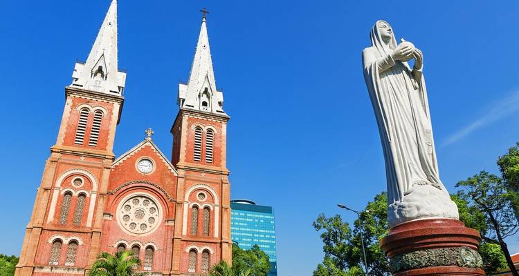 La Basilique-Cathédrale Notre-Dame de Saigon aux briques rouges s'élève majestueusement dans un ciel bleu clair à côté d'une statue blanche de la Vierge Marie.