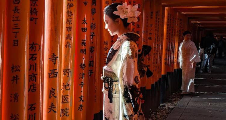 Woman in traditional attire standing among orange Torii gates with Japanese inscriptions.