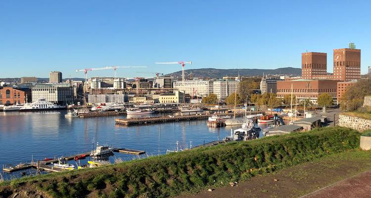 Le port en bord de mer d'Oslo avec des grues, des bateaux et l'hôtel de ville en briques rouges sous un ciel bleu dégagé.