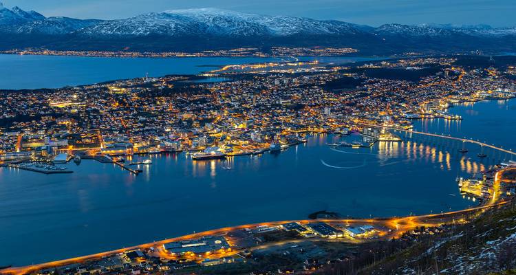Panorama nocturne de Tromsø illuminée aux côtés de fjords sombres et de montagnes saupoudrées de neige.