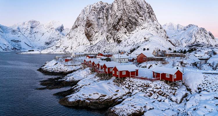 Des cabanes rouges couvertes de neige longent un rivage rocheux dominé par des pics dentelés spectaculaires à l'aube.