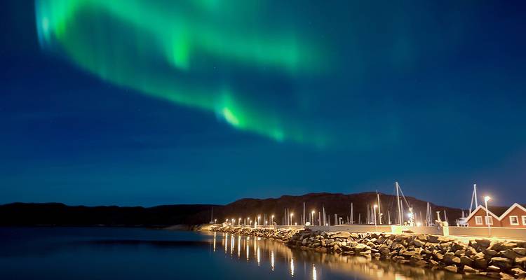 Une aurore boréale d'un vert éclatant s'étend en arc de cercle dans un ciel nocturne bleu profond au-dessus d'une promenade portuaire tranquille.