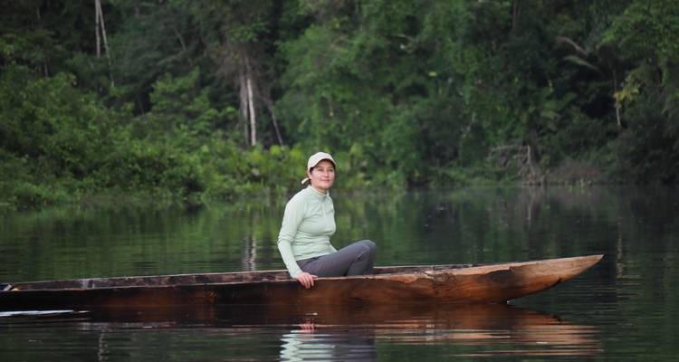 Une femme vêtue d'un haut vert clair est assise dans une pirogue en bois sculptée sur un fleuve de jungle aux eaux parfaitement immobiles.