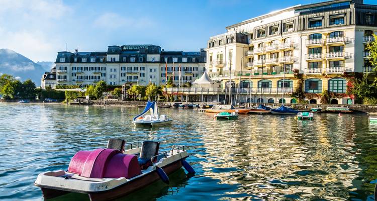 Seeblick mit Tretbooten und großem Hotelgebäude, das sich im Wasser spiegelt.