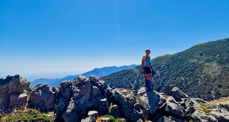 Ein Einzelwanderer steht auf zerklüfteten Felsen und blickt über bewaldete korsische Berge unter einem klaren blauen Himmel.