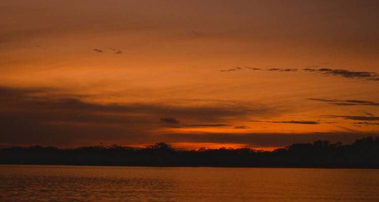 A fiery orange sunset reflects on calm river water with a dark treeline silhouette.