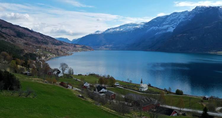 Un tranquilo pueblo junto al lago con montañas nevadas que se alzan más allá de verdes pastos y casas de granja dispersas.